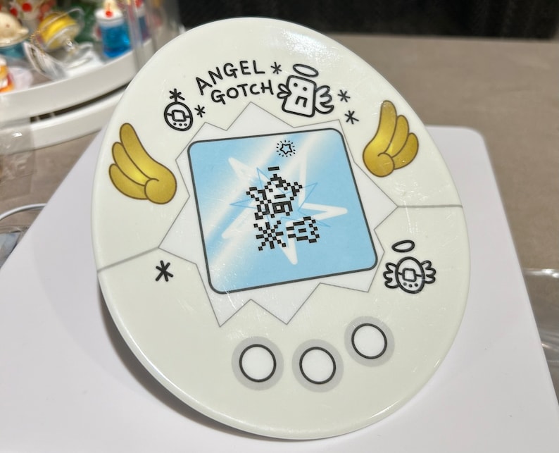 Pu&ograve; includere: Videogioco elettronico portatile bianco con la scritta "ANGEL GOTCH" e una grafica pixelata sullo schermo. Il dispositivo presenta ali d'angelo color oro e una piccola illustrazione di angelo. Tre pulsanti circolari si trovano in basso.