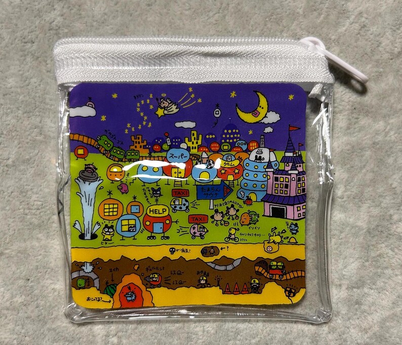 Pu&ograve; includere: Un astuccio quadrato trasparente con cerniera e un'illustrazione a cartoni animati colorata. L'illustrazione presenta un paesaggio urbano stravagante con edifici, veicoli e personaggi sotto un cielo stellato con una luna a mezzaluna. L'astuccio ha una cerniera bianca.
