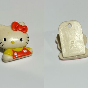 Pu&ograve; includere: Piccola statuina di Hello Kitty bianco sporco con fiocco rosso e completo giallo e rosso. La statuina ha occhi neri e baffi. Il retro ha una base rettangolare con un foro e la scritta "83 SANRIO".