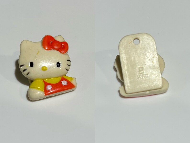 Pu&ograve; includere: Piccola statuina di Hello Kitty bianco sporco con fiocco rosso e completo giallo e rosso. La statuina ha occhi neri e baffi. Il retro ha una base rettangolare con un foro e la scritta "83 SANRIO".