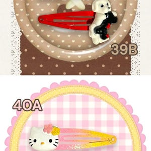 May include: A collection of hair clips featuring various designs. Includes blue dolphin clips, a house and dog clip, and Hello Kitty clips. The clips are on yellow and pink barrettes, with decorative backgrounds.