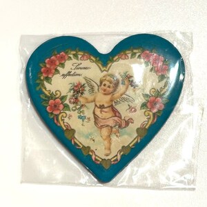 Pu&ograve; includere: Oggetto decorativo a forma di cuore con bordo turchese e immagine centrale di un cherubino che tiene dei fiori. L'immagine &egrave; circondata da rose rosa e le parole "Sincere affections". L'oggetto &egrave; in un involucro di plastica trasparente.