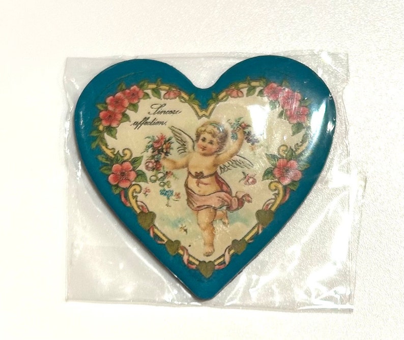 Pu&ograve; includere: Oggetto decorativo a forma di cuore con bordo turchese e immagine centrale di un cherubino che tiene dei fiori. L'immagine &egrave; circondata da rose rosa e le parole "Sincere affections". L'oggetto &egrave; in un involucro di plastica trasparente.