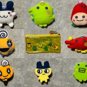 May include: A collection of colorful plush toys and accessories featuring cartoon characters. Includes a white and blue bunny, a green creature, a red strawberry, a yellow coin purse with text, and other character-shaped items.
