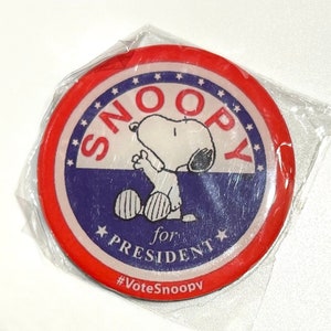 Pu&ograve; includere: Un bottone rotondo di Snoopy for President con un design rosso e blu. Il bottone presenta Snoopy in posa seduta, con la scritta "SNOOPY for PRESIDENT" e "#VoteSnoopy". Il bottone &egrave; in un involucro di plastica trasparente.