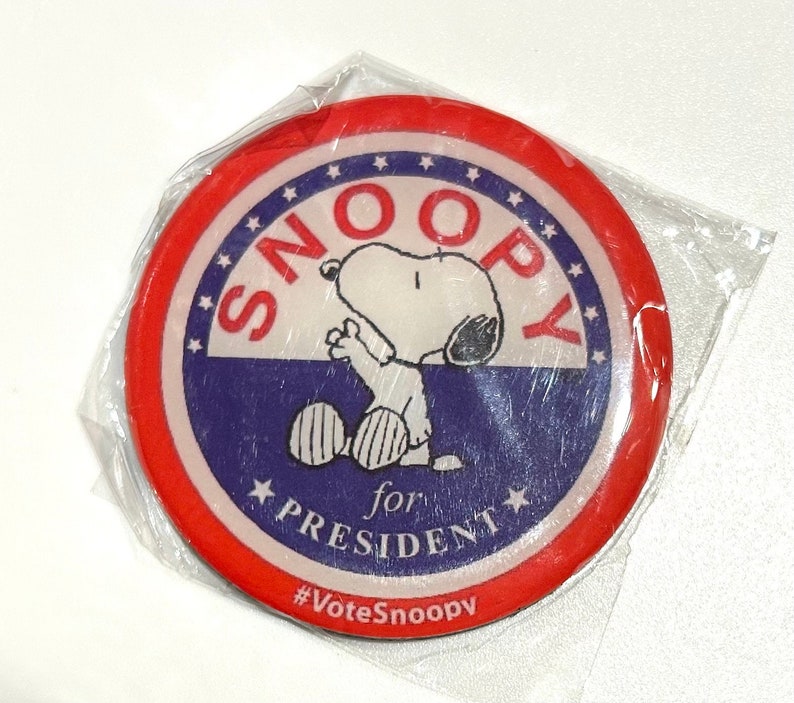 Pu&ograve; includere: Un bottone rotondo di Snoopy for President con un design rosso e blu. Il bottone presenta Snoopy in posa seduta, con la scritta "SNOOPY for PRESIDENT" e "#VoteSnoopy". Il bottone &egrave; in un involucro di plastica trasparente.