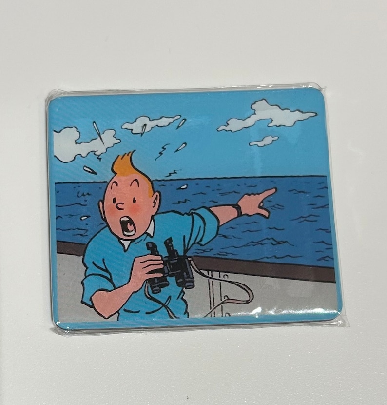 Pu&ograve; includere: Un sottobicchiere rettangolare con un'illustrazione a cartoni animati di Tintin, con la bocca aperta per la sorpresa, che indica il mare mentre tiene un binocolo. Lo sfondo mostra un cielo blu con nuvole e onde.