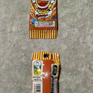 Pu&ograve; includere: Una confezione di snack giapponesi Umaibo. La confezione arancione e gialla presenta un personaggio dei cartoni animati e testo giapponese. Lo snack &egrave; un soffio di mais cilindrico.