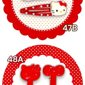 May include: A collection of hair accessories featuring cartoon character designs. Includes brown and white Hello Kitty clips, red polka dot clips, red fork-shaped clips, and a pink strawberry clip.
