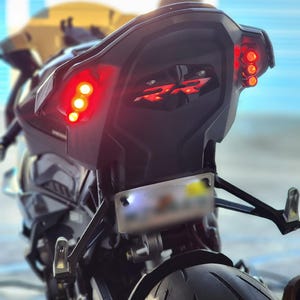 May include: Black motorcycle with bright red tail lights and a license plate. The motorcycle has a sleek, modern design with a black finish. The tail lights are illuminated, and the motorcycle appears to be in motion.