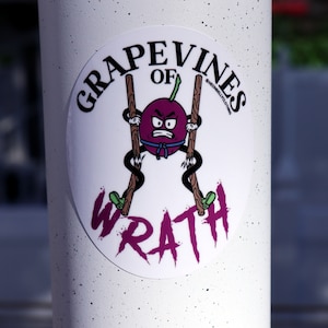 May include: A white sticker with a purple cartoon grape character holding two wooden sticks. The text "Grapevines of Wrath" is written in a pink, stylized font around the character.