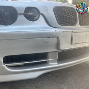 Può includere: Parte anteriore di un'auto argentata con griglie a nido d'ape nere e fari. Il paraurti ha una presa d'aria inferiore e un'area per la targa. Il logo "Printastic Workshop" è visibile in alto a destra.