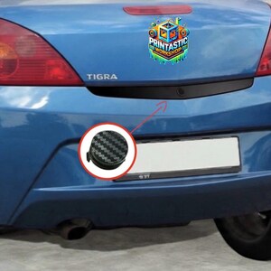 Peut inclure: Arrière d'une voiture bleue avec un pare-chocs noir et un autocollant "Printastic Workshop". Un gros plan montre un objet noir et gris à motif fibre de carbone. Le nom du modèle de la voiture, "TIGRA", est visible.