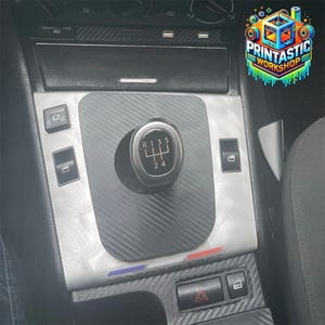 Op de afbeelding: Een middenconsole-afdekplaat voor een auto met een zilverkleurige geborstelde metalen afwerking en een centraal inlegstuk met zwarte koolstofvezeltextuur. De versnellingspookknop toont een standaard handgeschakeld patroon met vijf versnellingen.