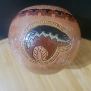 May include: A round, tan-colored ceramic vessel featuring a carved and painted bear motif in dark brown, black, and light blue. The upper section is decorated with a geometric band in dark red and black, with etched details throughout the surface.