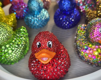 May include: A collection of colorful rubber duck toys covered in sparkling rhinestones. The ducks are arranged in a circle on a white surface. One duck is red with a bright orange beak and pink mouth.