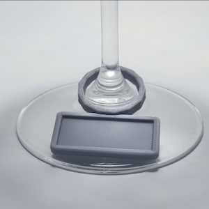 Puede incluir: Un charm para copa de vino gris con una etiqueta rectangular adjunta. El charm está diseñado para ajustarse alrededor del tallo de una copa de vino.