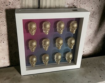 May include: A white frame with a gradient pink and blue background. The frame contains 15 gold glitter skull figurines.