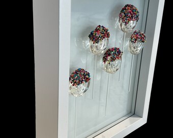 May include: A white framed shadow box with six glass spheres covered in colorful sprinkles. The spheres are attached to clear glass stems and are arranged in a cluster.