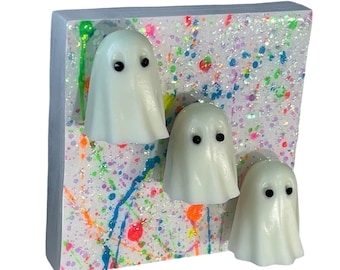 May include: Three white ghost figurines with black eyes are attached to a small wooden square. The square is covered in white glitter and has colorful paint splatters.