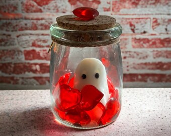 May include: A clear glass jar with a cork lid and a white ghost figurine inside. The jar is filled with red heart-shaped candies.