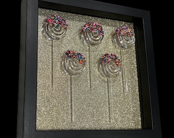 May include: A framed artwork featuring five glass lollipops with colorful sprinkles. The lollipops are arranged in a row and are set against a silver glitter background.