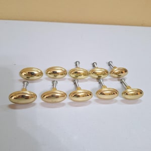 May include: A collection of ten gold-colored oval cabinet knobs with silver stems. The knobs are arranged in two rows, showcasing their polished, reflective surfaces. These hardware pieces are designed for furniture.