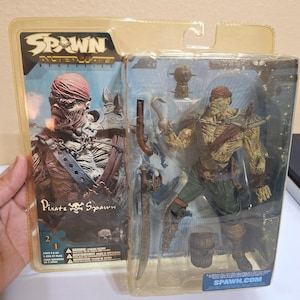 Peut inclure: Figurine d'action Pirate Spawn dans un emballage plastique. La figurine est un pirate zombie avec un crochet, une épée et un pistolet. L'emballage porte l'inscription "Spawn Alterate Realities" et "Pirate Spawn".