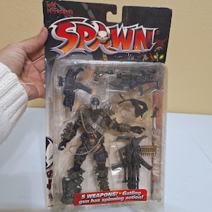 Peut inclure: Figurine de Spawn, un personnage de bande dessinée, dans un emballage plastique. La figurine est sombre avec un masque en forme de crâne, une armure à pointes et une chaîne. L'emballage comprend 5 armes, dont une mitrailleuse Gatling. Le texte "Todd McFarlane's Spawn" est en haut.
