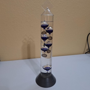 May include: A Galileo thermometer with a clear glass cylinder and a dark grey base. The cylinder contains several blue glass spheres with numbered tags. The thermometer is a decorative item used to measure temperature.