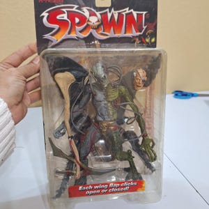 May include: Spawn action figure in a clear plastic package. The figure is a detailed, multi-colored character with wings and a skeletal appearance. The packaging includes the Spawn logo and the text "Each wing flap clicks open or closed!"