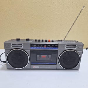 Op de afbeelding: Een grijze draagbare stereocassetterecorder met twee ronde zwarte luidsprekers, een centraal cassettedeck en een lange zilveren telescopische antenne. Het voorpaneel bevat een radiofrequentiedisplay met FM- en AM-banden, knoppen en het Samsung-logo.