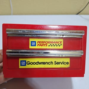 May include: A red metal storage chest with two drawers, each featuring a chrome handle. The top drawer has a yellow sticker reading GM PERFORMANCE PARTS with a checkered flag design. The bottom drawer has a yellow sticker reading GM Goodwrench Service.