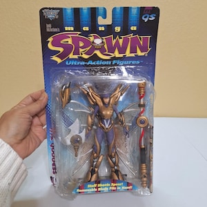 Puede incluir: Una figura de acción empaquetada del personaje "Spawn" de la serie "Ultra-Action Figures". La figura es un personaje femenino con armadura dorada y morada, alas y accesorios. El embalaje incluye el texto "Spawn" y "Staff Shoots Spear!"