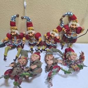 May include: A collection of eight decorative elf figurines featuring pointed hats with blue and white polka dots, red fabric accents, and detailed facial features. The figures are dressed in varied textured clothing, including striped pants and vests, with some holding small decorative greenery.