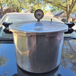 Könnte beinhalten: Ein silberner Schnellkochtopf mit schwarzem Griff und Manometer oben. Der Kochtopf steht auf der Motorhaube eines Autos, im Hintergrund Bäume und Häuser. Das Manometer hat ein schwarzes Zifferblatt mit weißen Zahlen und einer Nadel.