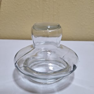 May include: A clear glass bottle stopper with a rounded top and a flared base. The stopper has a unique shape, with a bulbous body and a narrower neck. The glass is transparent, allowing light to pass through.