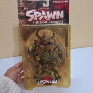 Puede incluir: Una figura de acción de McFarlane Toys de la serie Dark Ages Spawn, "The Samurai Wars". La figura está en un embalaje de plástico transparente. La figura es un guerrero detallado con un casco con cuernos, armadura y un arma. El embalaje incluye el sitio web spawn.com.