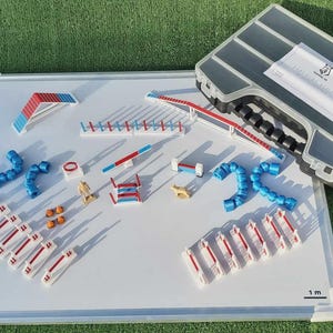 Peut inclure: Un tableau blanc magnétique marqué TANIERE 3D avec une échelle de 1m, présentant divers obstacles miniatures d'agilité canine en rouge, blanc et bleu, incluant des sauts, tunnels et une bascule, avec une mallette de rangement étiquetée.