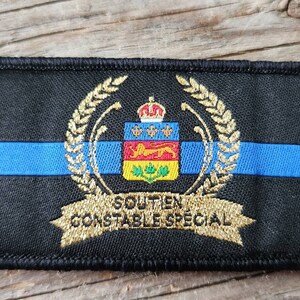 Peut inclure: Écusson noir brodé avec une bande bleue et des détails dorés. Il présente un écusson avec une couronne, des armoiries et le texte "SOUTIEN CONSTABLE SPECIAL" en or. De forme rectangulaire.