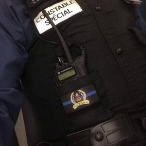 K&ouml;nnte beinhalten: Eine schwarze Weste mit dem Text "CONSTABLE SP&Eacute;CIAL" auf einem wei&szlig;en Aufn&auml;her. Ein Motorola-Funkger&auml;t befindet sich in einer Tasche, und ein Aufn&auml;her mit blauer Linie und Emblem ist sichtbar.