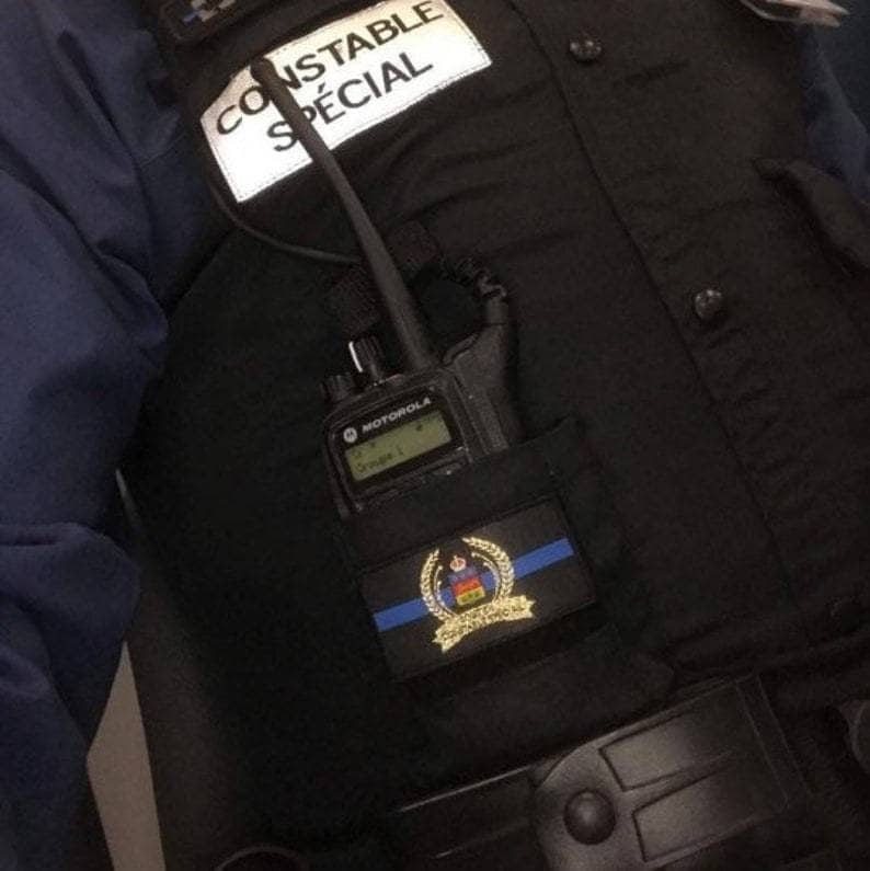K&ouml;nnte beinhalten: Eine schwarze Weste mit dem Text "CONSTABLE SP&Eacute;CIAL" auf einem wei&szlig;en Aufn&auml;her. Ein Motorola-Funkger&auml;t befindet sich in einer Tasche, und ein Aufn&auml;her mit blauer Linie und Emblem ist sichtbar.