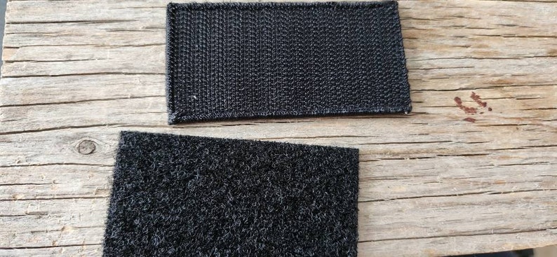 K&ouml;nnte beinhalten: Zwei schwarze rechteckige Klettverschluss-Patches. Einer hat eine gewebte Textur, der andere eine flauschige Textur. Die Patches werden auf einer verwitterten Holzoberfl&auml;che gezeigt.
