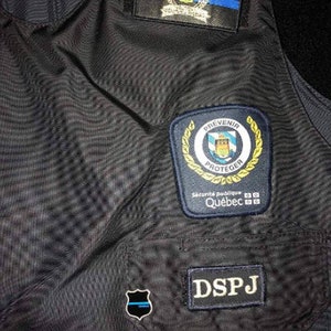 K&ouml;nnte beinhalten: Nahaufnahme einer dunkelblauen Weste mit mehreren Aufn&auml;hern. Zu den Aufn&auml;hern geh&ouml;ren ein Wappen mit einer blauen Linie, ein Aufn&auml;her mit den Worten "PREVENIR PROTEGER" und "Qu&eacute;bec" sowie ein Aufn&auml;her mit der Aufschrift "DSPJ".