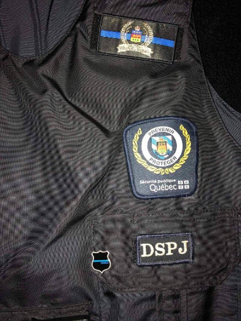 K&ouml;nnte beinhalten: Nahaufnahme einer dunkelblauen Weste mit mehreren Aufn&auml;hern. Zu den Aufn&auml;hern geh&ouml;ren ein Wappen mit einer blauen Linie, ein Aufn&auml;her mit den Worten "PREVENIR PROTEGER" und "Qu&eacute;bec" sowie ein Aufn&auml;her mit der Aufschrift "DSPJ".