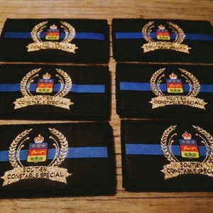 K&ouml;nnte beinhalten: Sechs schwarze, rechteckige Aufn&auml;her mit blauem Streifen und goldenem Emblem. Das Emblem zeigt eine Krone, ein Schild mit farbenfrohem Design und die Worte "SOUTIEN CONSTABLE SPECIAL".