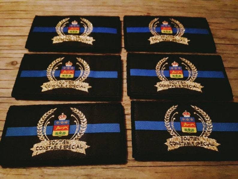 K&ouml;nnte beinhalten: Sechs schwarze, rechteckige Aufn&auml;her mit blauem Streifen und goldenem Emblem. Das Emblem zeigt eine Krone, ein Schild mit farbenfrohem Design und die Worte "SOUTIEN CONSTABLE SPECIAL".