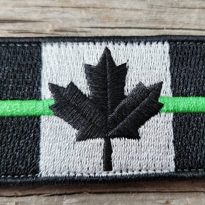 Peut inclure: Écusson brodé avec un motif de drapeau canadien stylisé. Le patch a une bordure noire, un fond rayé noir et blanc, une feuille d'érable noire et une bande verte horizontale. De forme rectangulaire.