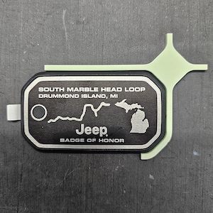 May include: Black and silver Jeep Badge of Honor tag with the text "South Marble Head Loop Drummond Island, MI" and an outline of the state of Michigan. The tag has a silver Jeep logo and the text "Badge of Honor".