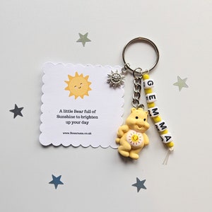 May include: A yellow bear keyring with a smiling sun face and the words "A little Bear full of Sunshine to brighten up your day" on a white card. The keyring also has a silver charm with a sun design and yellow beads spelling "GEMMA".